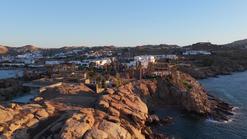 Mykonos island in Greece flying over iconic whitewashed houses and rocky coast during a warm summer sunset over Aegean Sea with calm blue water and clear skies