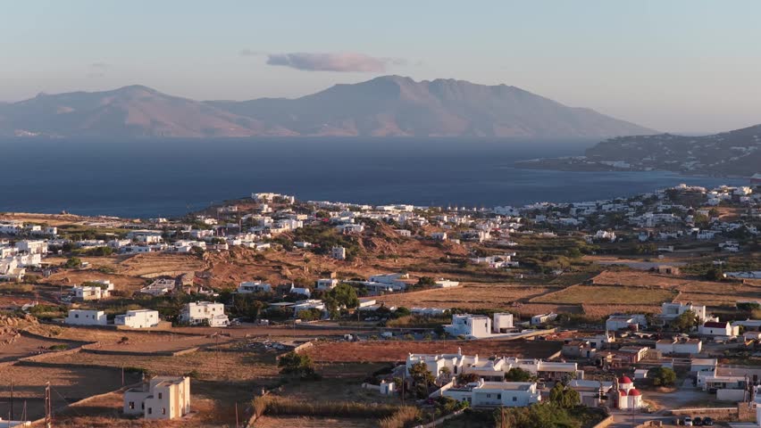 Mykonos whitewashed villages dotting arid Cycladic landscape with Aegean Sea and mountainous Tinos island during summer golden hour from tripod elevated perspective