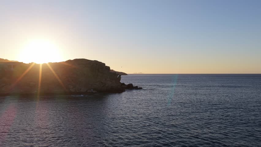 Golden sunlight reflecting over calm Aegean Sea near rocky coast of Mykonos, Greece during a summer sunset, Mediterranean travel and vacation scenery
