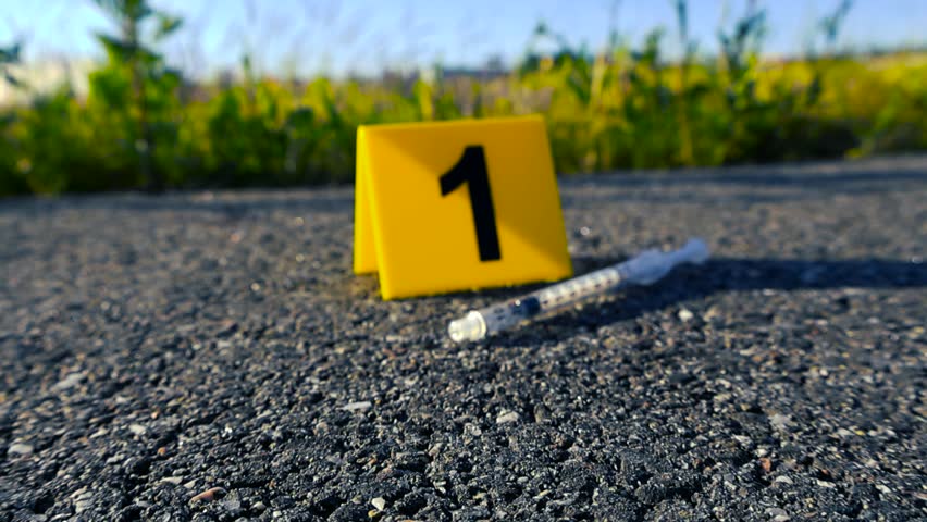 Close up view of a yellow evidence marker with the number one and a used syringe on an asphalt road at a crime scene, suggesting a police investigation related to drug use or murder