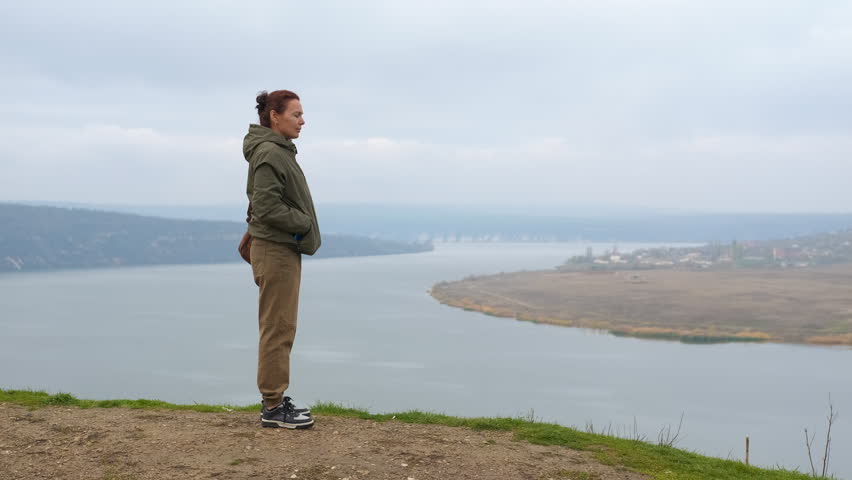 Woman standing on cliff edge contemplating the river view. Pensive woman standing alone on a scenic clifftop, enjoying the tranquil beauty of the river and landscape on a windy, cloudy day