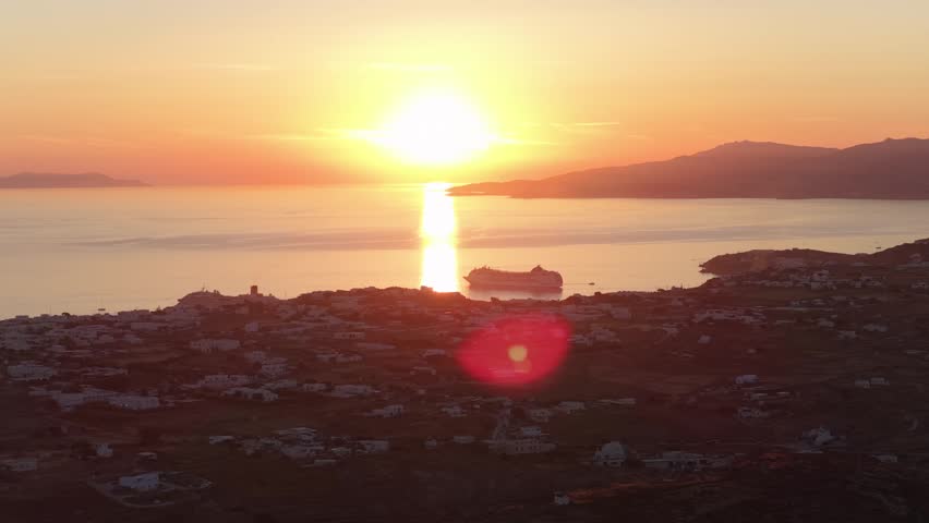 Golden summer sunset over Mykonos Town, Aegean Sea reflections, cruise ship in Tourlos bay, lens flare from setting sun, capturing Greek holiday mood