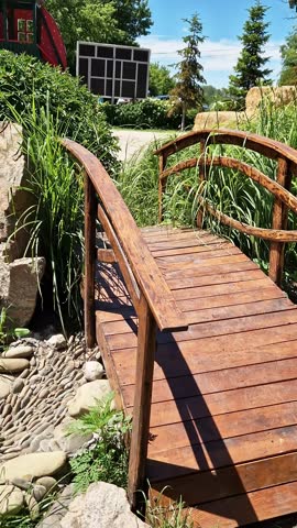 A rustic wooden arched bridge spans across a dry stream bed made of smooth river rocks and lush green garden grasses on a bright sunny day. Concept of landscaping and garden sanctuary.