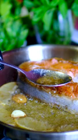Salmon steak basted with butter by chef using spoon while garlic and lemon cook in hot pan during seafood recipe close up