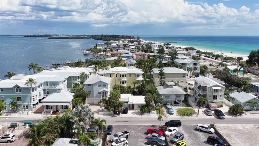 Colorful beach houses line coastal street, turquoise Gulf waters, sandy beach, palm trees, parked cars, summer clouds, distant island, Bradenton, Florida, United States. Aerial drone wide shot