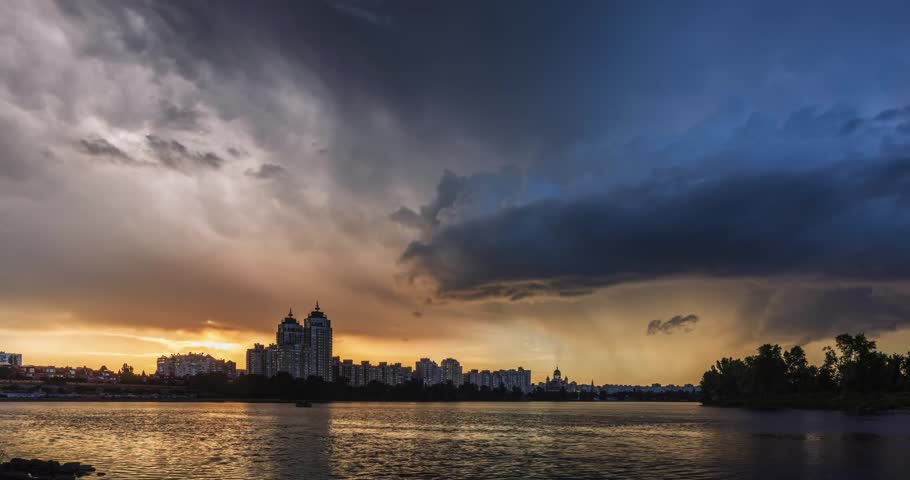 Dramatic sunset after a thunderstorm over Obolon Embankment in Kyiv, Ukraine. Golden sunbeams break through storm clouds, reflecting across the Dnipro River and city skyline.

