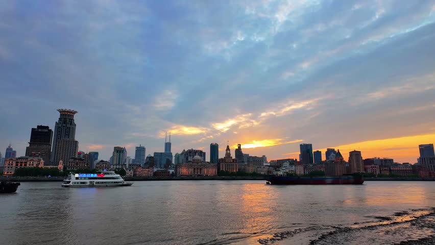 A riverfront urban skyline at dusk, with boats on calm water and sunset glow reflecting on the surface.