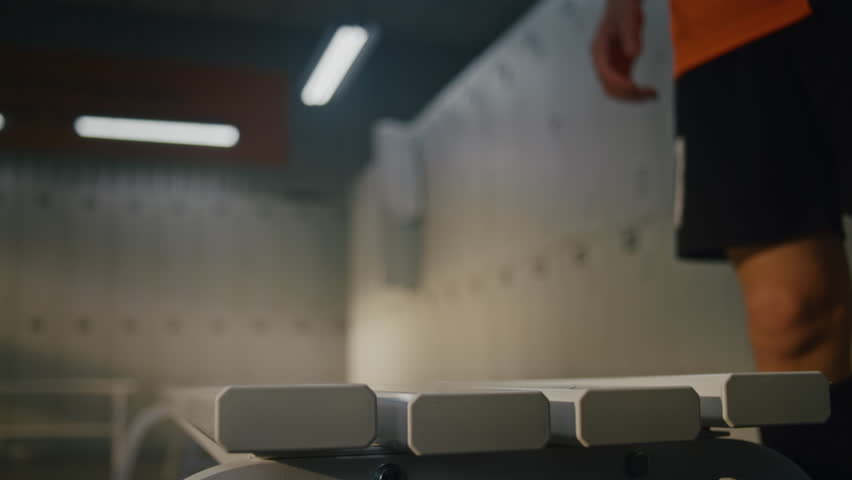 Young Footballer in Orange Jersey Preparing For Training Session in Locker Room. Boys Hands Methodically Ties Shoelaces of Athletic Cleats, Pulls Up Black Sports Socks. Focus Before Entering Field.
