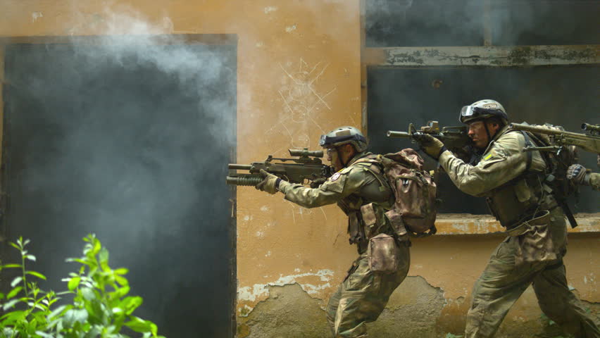 Ultra slow motion soldiers move tactically along a building wall, aiming rifles through drifting smoke.