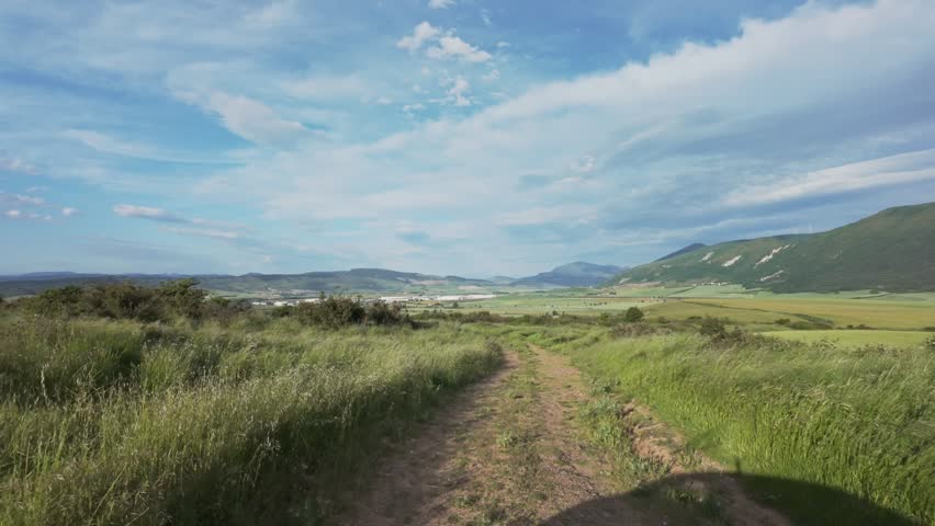 A scenic landscape with a dirt path winding through a grassy field, framed by rolling hills and distant mountains. The sky above is partly cloudy