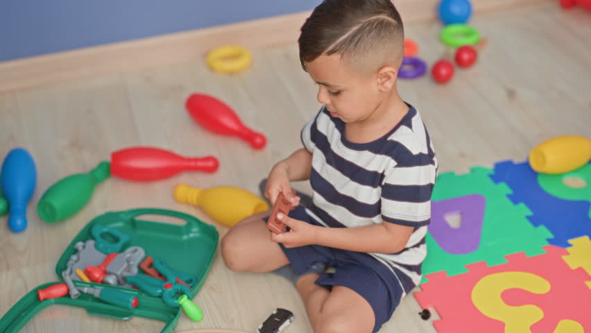 Little boy sitting on playroom floor holding toy toolkit pieces with striped shirt visible in studio; quiet concentration.