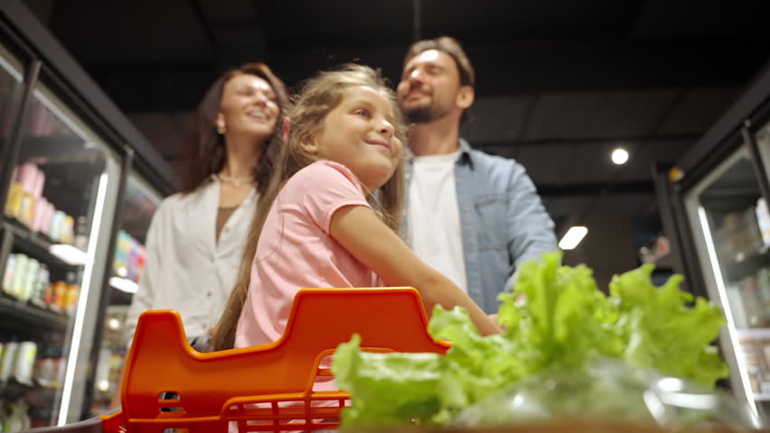 Smiling little girl sits calmly in shopping cart as parents walk along supermarket aisle. Family with preschooler daughter does shopping in grocery store
