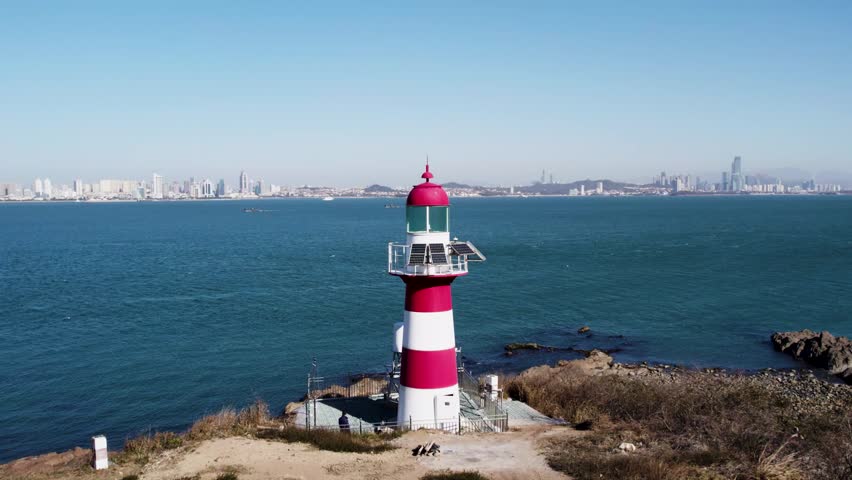 A red and white striped lighthouse stands on a coastal shore overlooking a blue sea, with a distant city skyline under a clear sky.