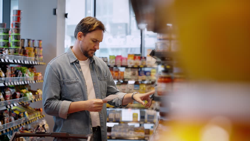 Focused man checks shopping list and takes tin can in grocery. Young bearded customer in denim seeks regular food product for eating in local supermarket