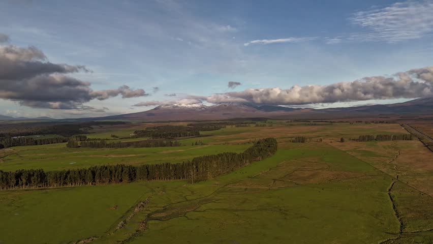 Motionlapse of clouds moving over distant snow capped peak of Tongariro mountain beyond green field at dusk in Tongariro National Park, New Zealand. Aerial forward