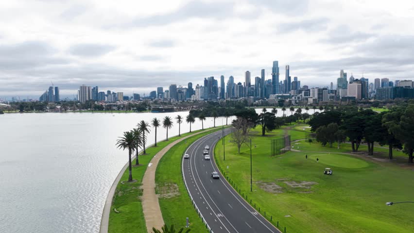 Beautiful cinematic aerial drone footage of Albert Park Lake featuring the curving lakeside road with moving traffic, cyclists, palm trees, expansive parklands, and the Melbourne CBD skyline backdrop