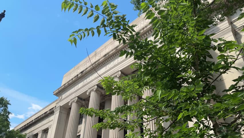 MIT Building 10 in Cambridge, Massachusetts, with its iconic neoclassical columns and stone facade framed by green tree branches under a clear blue sky. The image captures the historic academic archit