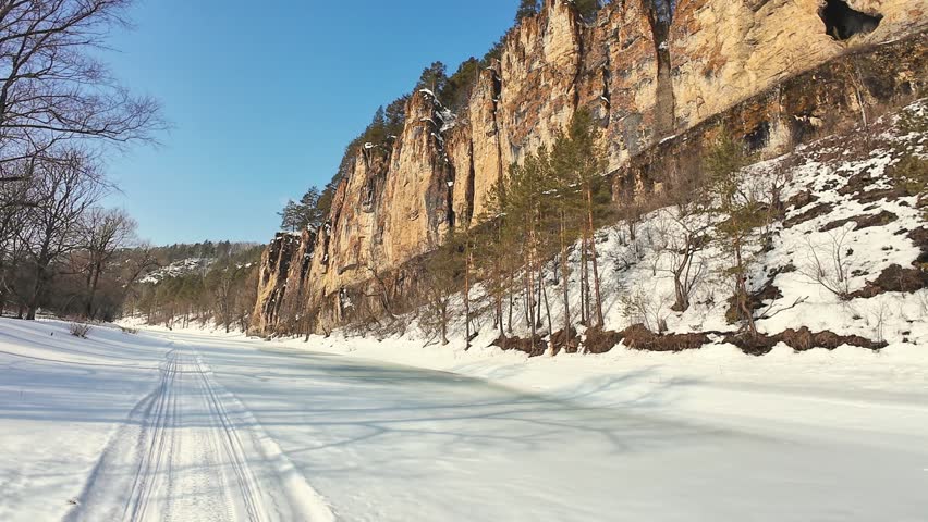 Frozen Waters Wind Through High Cliffs. Icy River Winds Past Snowy Pine Trees And Rocky Cliffs. Frosty Landscape With Flowing Icy Streams Amid Rugged Mountain Surroundings