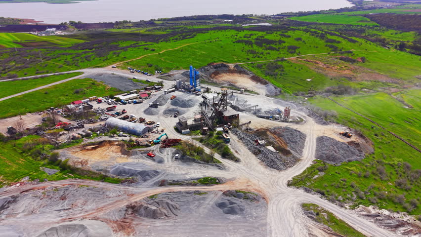 Quarry processing plant near lake. Elevated angle reveals silos, storage tower, gravel piles and surrounding green farmland.