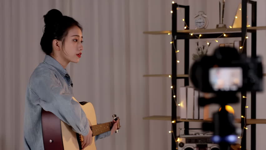 Talented girl musician looks at sheet music note for guitar on music stand, practicing playing acoustic guitar. Teacher recording online video course on camera playing acoustic guitar lesson tutorial