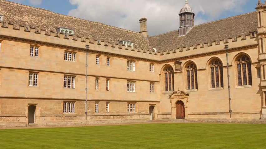 Historic stone college courtyard with arched windows and green lawn under sunlight