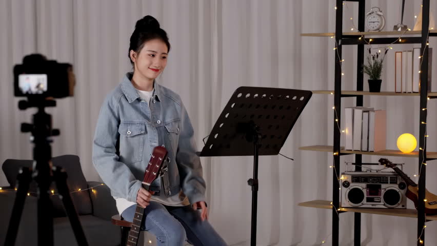 Musician looks at note for guitar on music stand, practicing playing acoustic guitar. Teacher reviews new material at sheet music in front of her while performing song with instrumental accompaniment