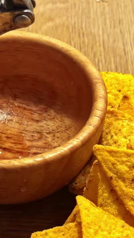 Garlic cloves are pressed with a metal garlic press into a wooden bowl placed beside crunchy nacho chips on a wooden surface.