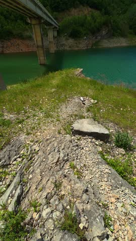 Lush green grass lines a rocky lakeshore. Teal water reflects a bright blue sky. Vertical video. Location: Zavoj Lake (Zavojsko jezero), Balkan Mountains (Stara Planina). Balkans, Serbia.