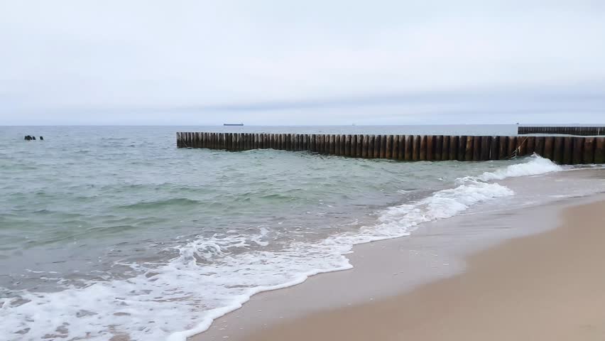 Tranquil seaside landscape with stormy coastal surf water waves rolling on empty sand beach and old wood breakwater on Baltic sea shore under cloudy sky. No people seascape natural background.