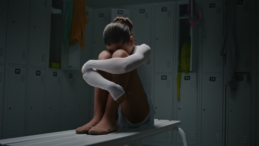 Young Female Athlete Sitting Isolated on Long Bench inside Dark, Empty Dressing Room. Dressed in White Leotard, Girl Processing Deep Emotional Pain of Lost Competition, Failure, and Heavy Self Doubt.