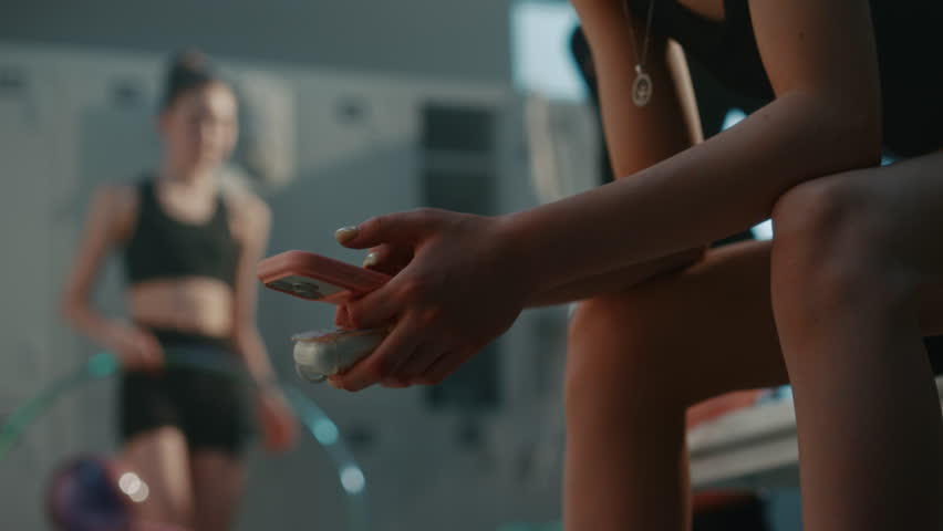 Close Up of Young Female Athlete Hands as Sits on Gym Bench. Dressed in Black Activewear, Girl Holds Pink Smartphone and Case For Wireless Bluetooth Earbuds. Interaction With Modern Technology.