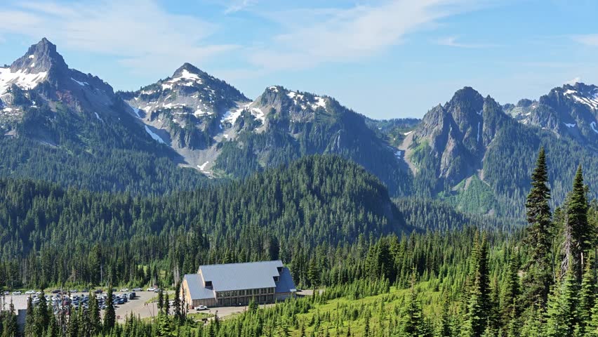 A wide shot of a large lodge with a grey roof and a busy parking lot surrounded by dense evergreen forest and snow-capped mountains in Washington state.