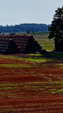 Stacked hay bales in pyramid shape on farmland beside open rural fields, drone, vertical farm industrial backdrop