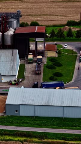 Trucks Parked Outside An Industrial Agricultural Silo And Storage Facility Next To Farmland Fields. vertical, aerial sideways shot