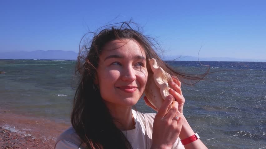 Happy young woman listening to the sound of the ocean in a seashell on a sunny beach