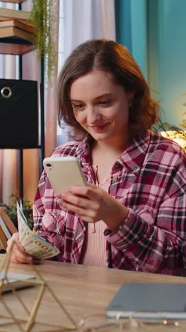 Rich excited Caucasian young woman freelancer looks at camera holding money cash and smartphone. Female businesswoman girl satisfied of income earnings saves money sitting at home office at table.