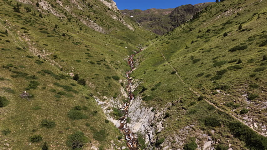 Aerial pullback reveals a cascading alpine stream flowing over iron-rich reddish rocks through a narrow green valley in Andorra, with snowy peaks, rocky slopes and lush meadows