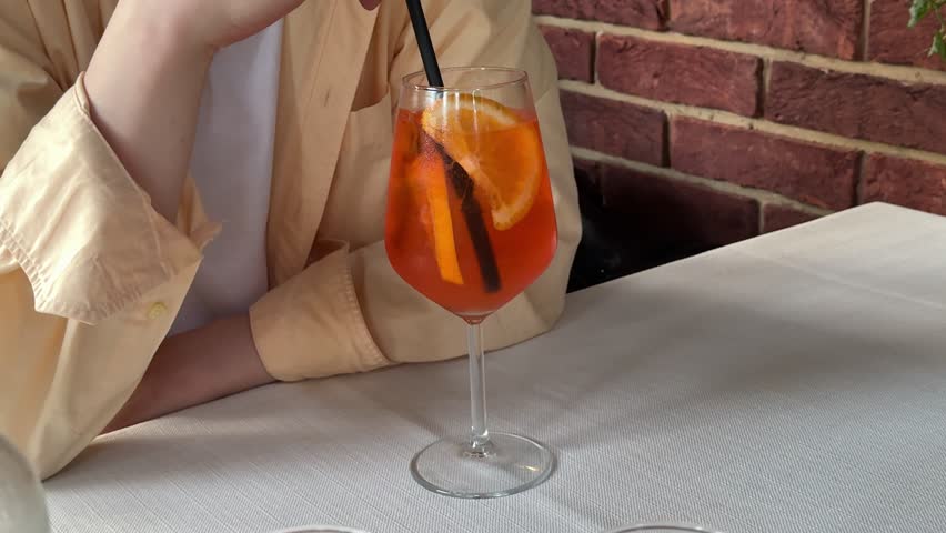 Unrecognizable man in yellow shirt stirring Aperol Spritz with black straw on elegant restaurant table with white tablecloth during relaxing dining experience