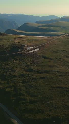 Vertical Screen: Beartooth Highway cuts through vast Montana USA alpine terrain with snow patches dotting green meadows and layered mountain ridges fading into hazy horizon at golden hour.