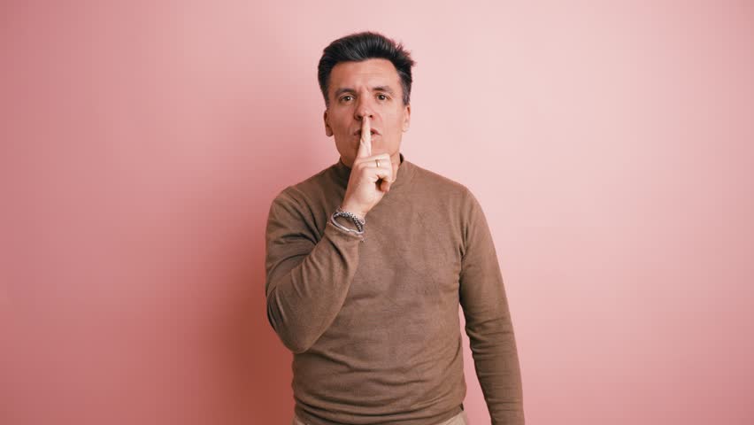 Handsome middle aged man looking at the camera with his index finger on his lips, asking for silence. Studio shot against a pink wall