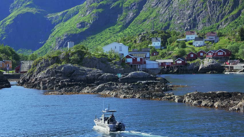 Small fishing vessel navigates crystal blue waters toward iconic red wooden houses built on stilts in dramatic Lofoten fjord, surrounded by