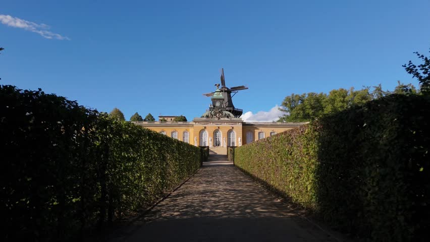The ornate facade of the New Palace rises beyond a symmetrical hedge-lined approach with an old windmill behind in Sanssouci Park, Potsdam.