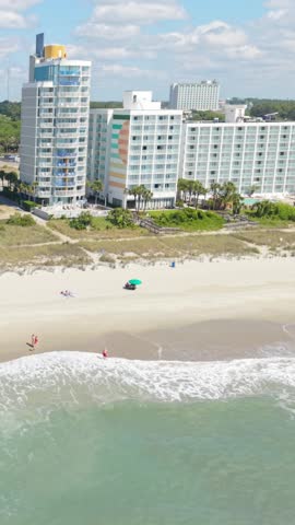 Vertical screen. Beachgoers enjoy sunny day at Myrtle Beach, South Carolina, with ocean waves crashing on sandy shore and hotels in the background