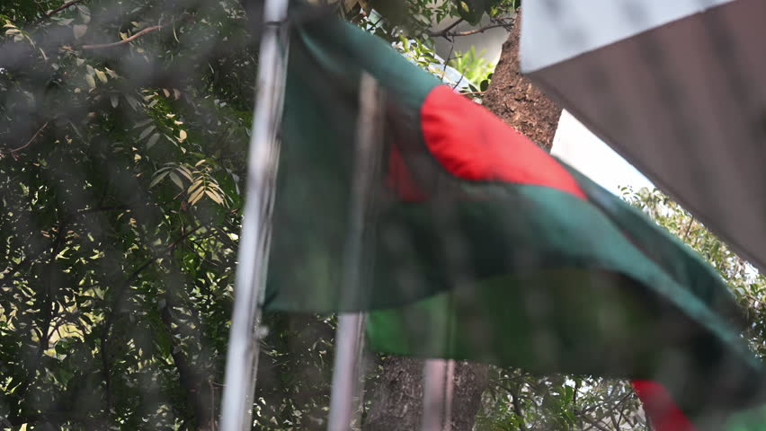The Bangladesh national flag and BNP party flag wave behind a blurred razor wire fence on a street in Dhaka. Captures themes of political tension, security enforcement, and regional election events.