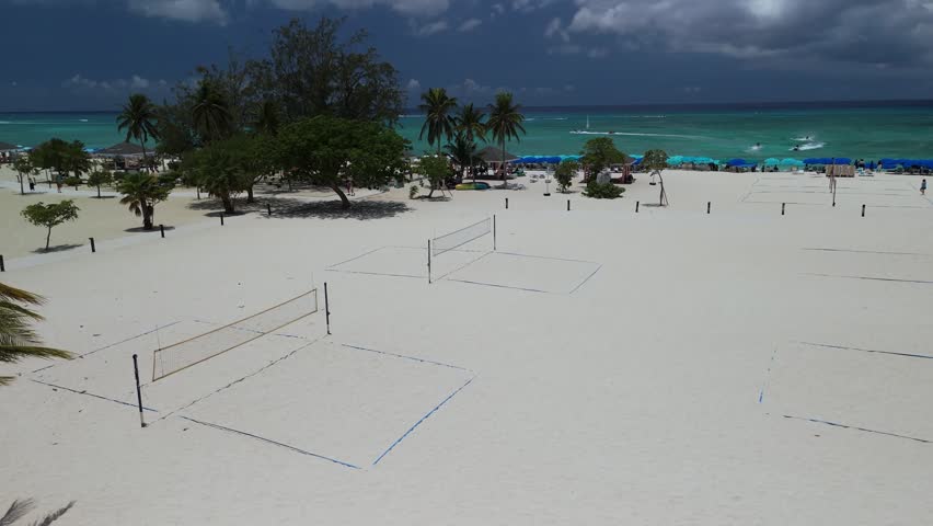 Aerial view of Grand Cayman beach with volleyball courts on white sand, turquoise water, and palm trees in the Cayman Islands.
