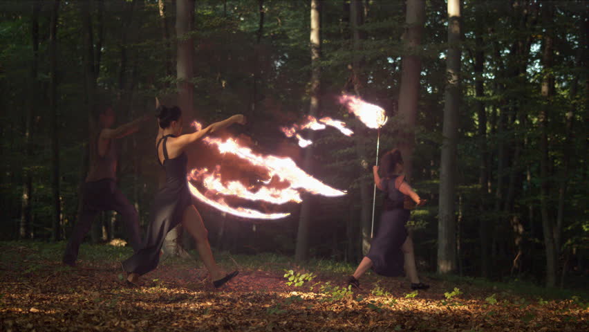 Artistic fire performance in a woodland setting, flames trailing behind performers in ultra slow motion.