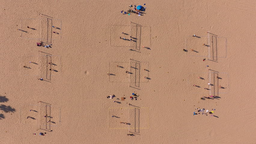 People playing volleyball on sandy beach courts in Santa Monica, California, USA. Drone top down view shows multiple volleyball nets with players casting long shadows on golden sand during sunny day.