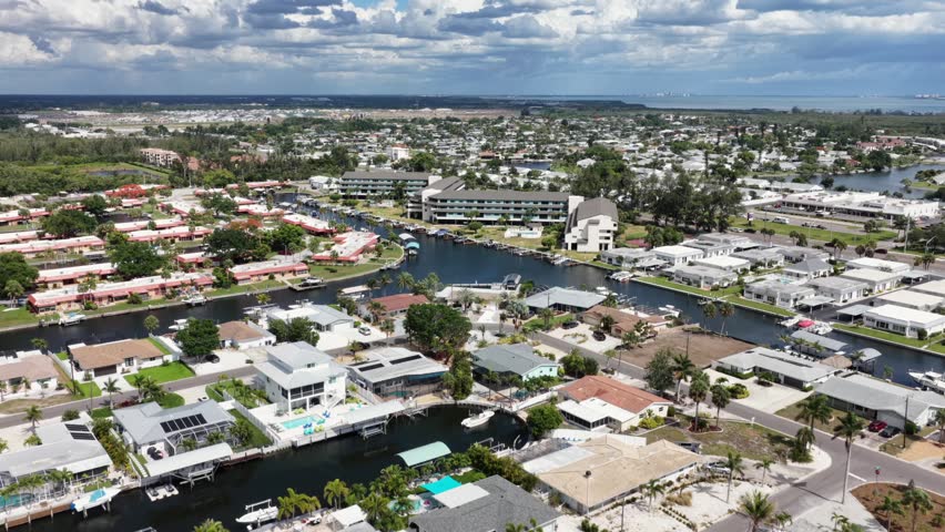 Canal community with waterfront homes and private boat docks winds through dense neighborhood, church tower, palm trees, cloudy sky, Bradenton, Florida, United States. Aerial drone wide shot