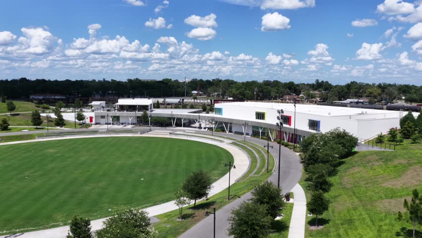 Bonnet Springs Park rolling green lawn curves toward modern pavilion buildings, winding path, scattered clouds, sunny blue sky, trees, Lakeland, Florida, United States. Aerial drone flyover shot