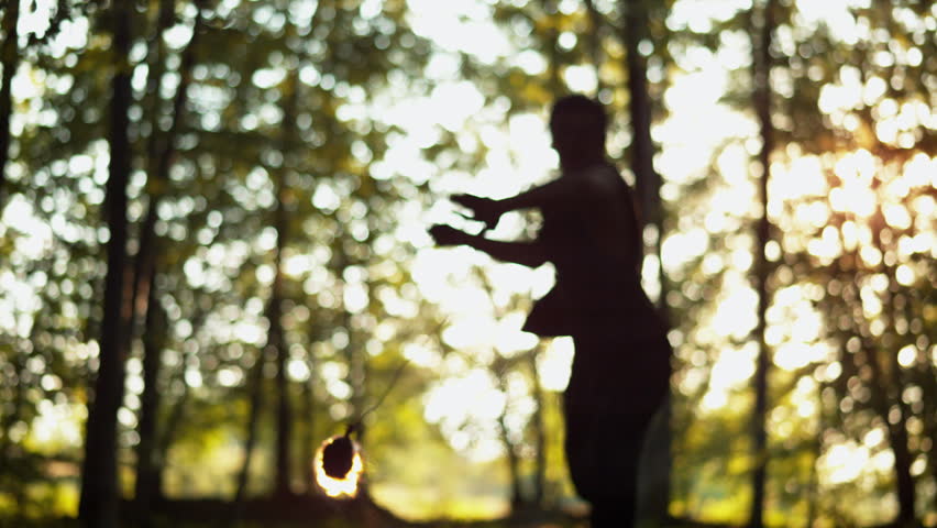 Backlit silhouette spins flaming poi in a wooded clearing, captured in ultra slow motion at golden hour.